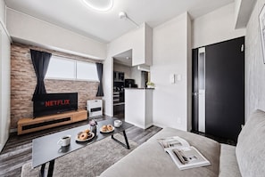 A modern living space with a flat-screen TV, a coffee table with pastries, a sofa, and a kitchen area visible through an open door. - J-STAR 802 (Sapporo)