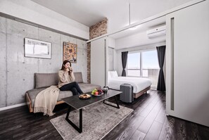 Una habitación de hotel moderna con sofá, cama, mesa de comedor con comida, maleta y vistas a la ciudad.