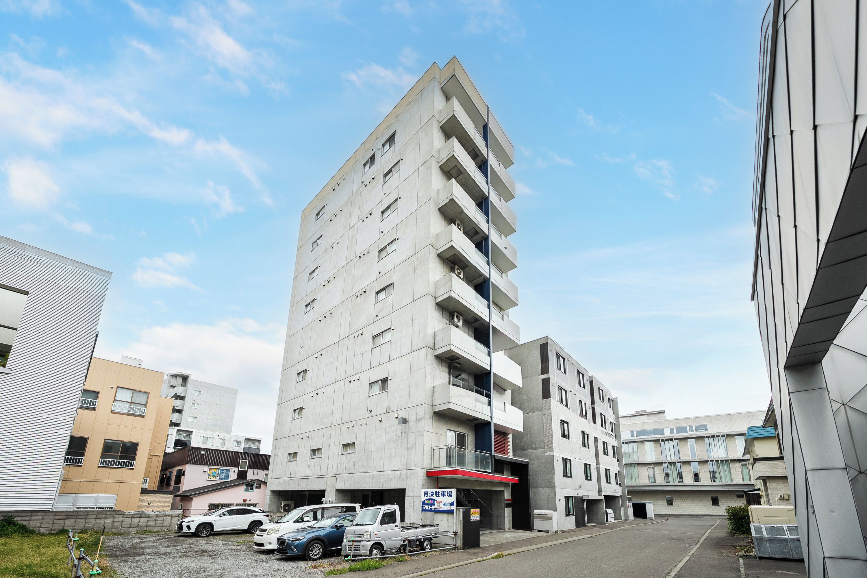 Un edificio de varios pisos con fachada plana, varios balcones y un estacionamiento con coches.