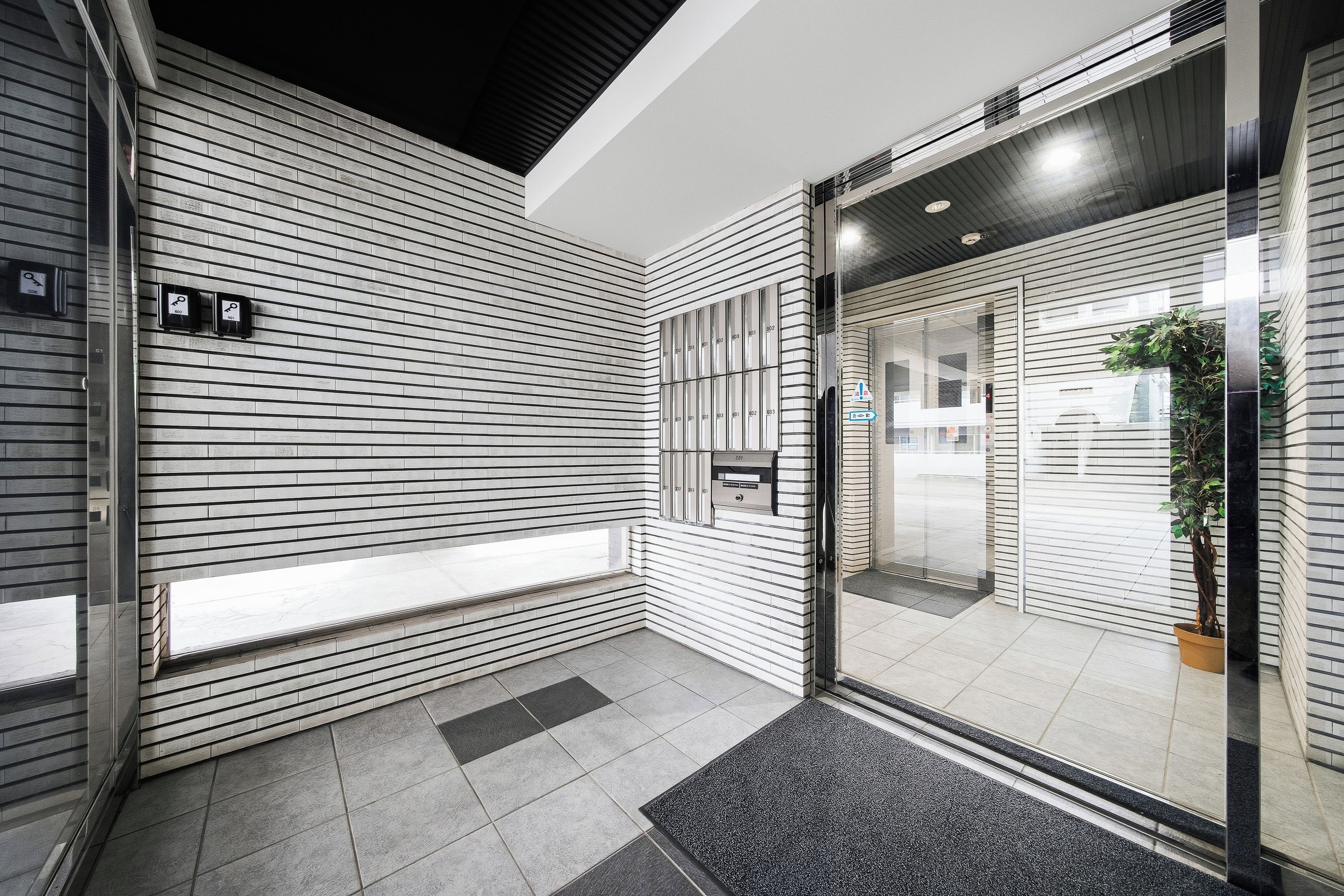 A modern building entrance with a glass door, a potted plant, and a tiled floor.