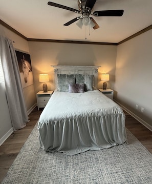 A bedroom with a bed, two bedside tables with lamps, a ceiling fan, and a large window with curtains. - Oxford Condo ~ Walk to Campus (Oxford)