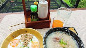 Desayuno con huevos y carne en un plato, un tazón de sopa y un vaso de jugo sobre una mesa con vista a un jardín y una fuente de agua.