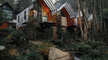 Un hotel moderno de estilo cabaña, ubicado en un área boscosa con un arroyo y grandes rocas.