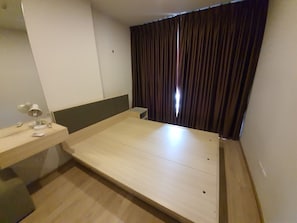A small, single-bed room with a wooden bed, a bedside table with a lamp, and a window with dark curtains. - 10 minutes walk to BTS bangna BITEC resort style condo 3 swimming pools 2 gyms (Krung Thep Maha Nakhon)