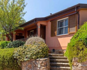 A house with a porch, steps leading to the entrance, and a stone wall with a number plaque. - Holiday home with a/c (Talamone)
