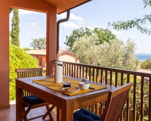 A wooden table with a yellow placemat, a white pitcher, and a bowl on a balcony with a wooden railing overlooking a landscape with trees and a building. - Holiday home with a/c (Talamone)