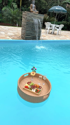 A pool with a floating basket containing food and drinks.