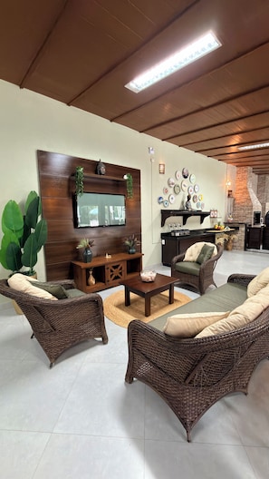 A living room with wicker furniture, a flat-screen TV, and a kitchen area with a brick oven.