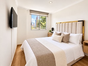 A modern bedroom with a large bed, a flat-screen TV mounted on the wall, a window with a view of buildings, and a nightstand with a lamp and a plant. - New Listing (Medellín)