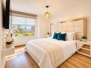 A modern bedroom with a large bed, a television mounted on the wall, a window with blinds, and a hanging light fixture. - New Listing (Medellín)