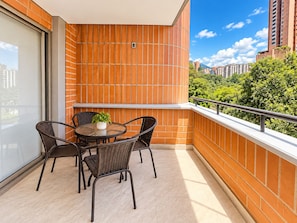 A balcony with wicker chairs and a small table, a potted plant, and a view of a cityscape with buildings and greenery. - New Listing (Medellín)