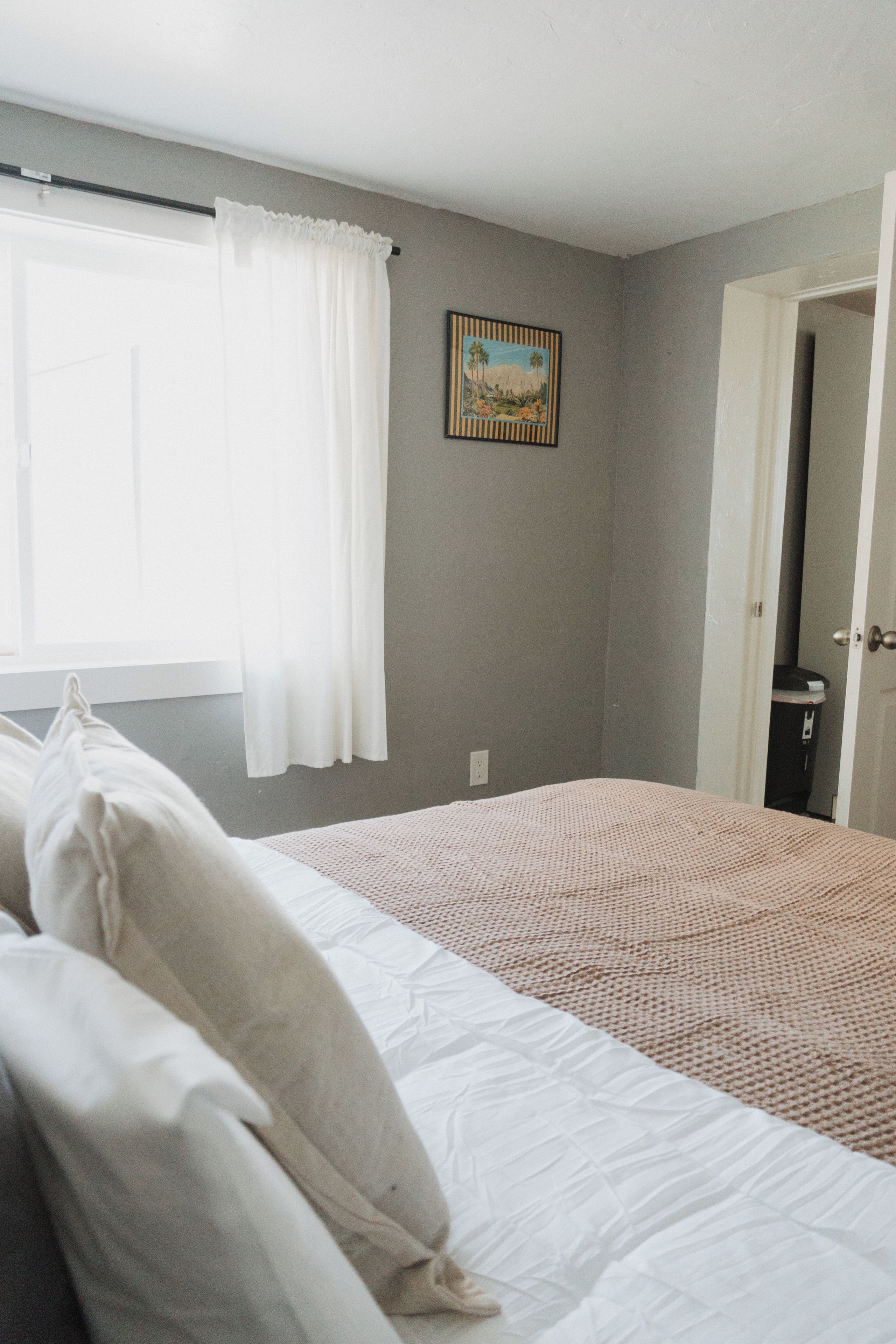 Quarto com cama, janela com cortinas, porta e quadro na parede.