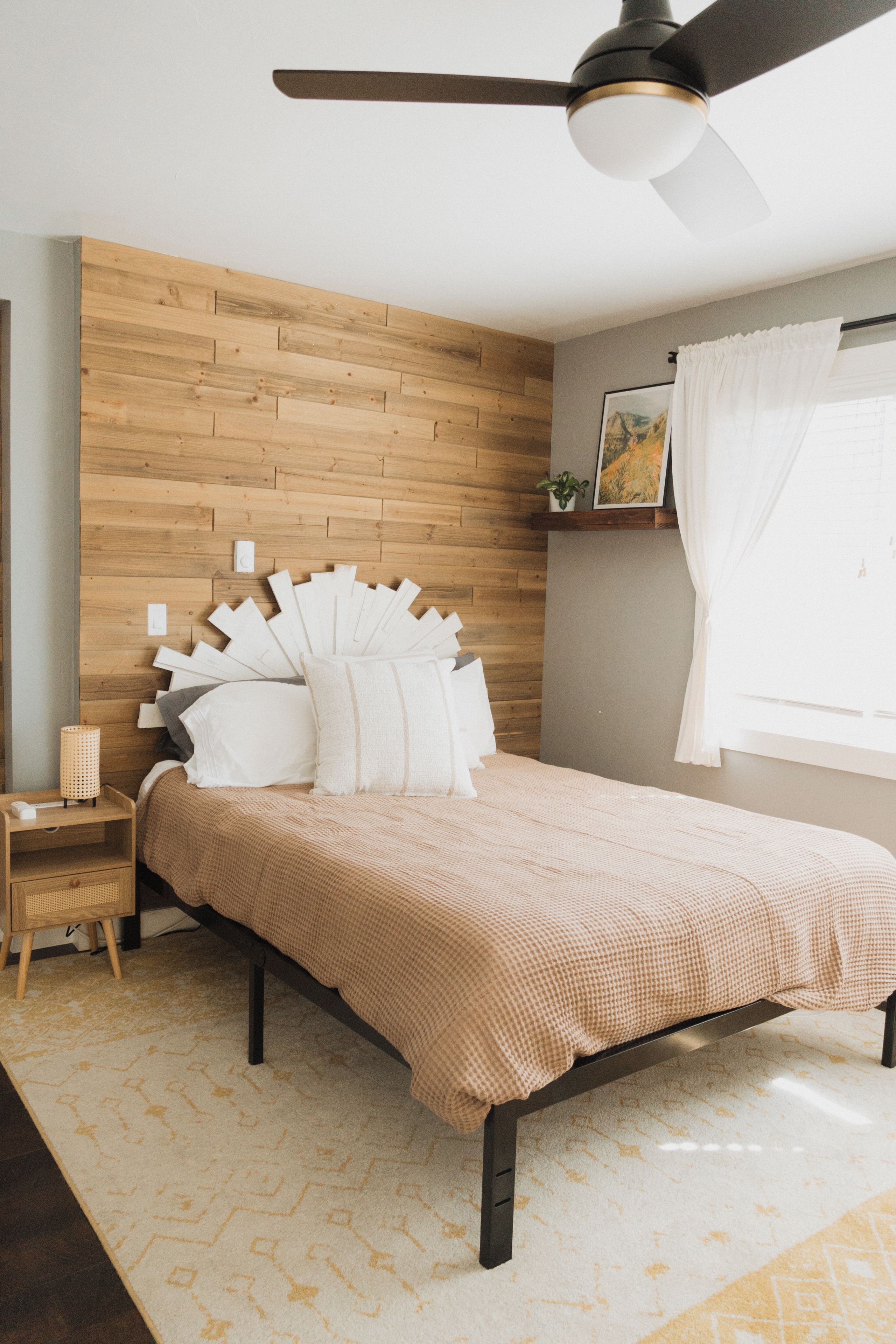 A bedroom with a wooden headboard, a bed with white and beige bedding, a nightstand with a lamp, and a ceiling fan.