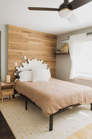A bedroom with a wooden headboard, a bed with white and beige bedding, a nightstand with a lamp, and a ceiling fan. - Enjoyable 3-bedroom house in Palmer Lake perfect for your getaway (Palmer Lake)