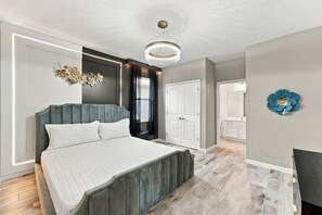 A modern bedroom with a large bed, a chandelier, a decorative wall piece, and a view of the bathroom. - Sonoma Resort | 15BR Mansion, Pool, Sleeps 30 (Kissimmee)