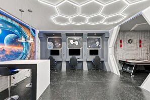 Una habitación con techo hexagonal, un escritorio con sillas y una pared decorada con motivos espaciales.