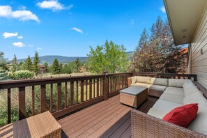 A deck with a sofa and a small table, surrounded by a wooden railing, overlooking a landscape with trees and distant mountains.