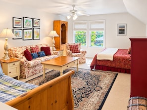 A bedroom with a bed, two chairs, a table, a wooden cabinet, a ceiling fan, and framed pictures on the wall. - South Main Street Cottage - Leland, Michigan - Saturday to Saturday (Leland)