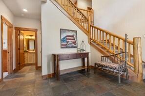 A staircase with wooden railings leading to an upper floor, a console table with drawers, a bench with a patterned cushion, and a wall-mounted American flag painting. - Trollhaugen Lodge | 4BR, Sleeps 10, Near Ski Base (Steamboat Springs)