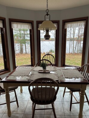 A dining table with placemats, chairs, and a hanging light fixture in a room with windows overlooking a body of water. - Cozy Cabin on a Private Point — Water Views on 3 Sides, Fishing & Trail Access (Irma)