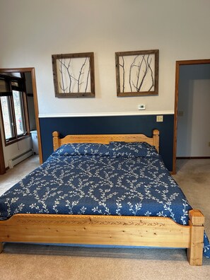 A bedroom with a wooden bed, a blue floral bedspread, two wall-mounted branch paintings, a thermostat, and a window with blinds. - Cozy Cabin on a Private Point — Water Views on 3 Sides, Fishing & Trail Access (Irma)