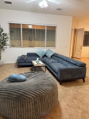 A living room with a sectional sofa, a round ottoman, a small table with a tissue box, and a potted plant. - Spacious 4-bedroom house in marvelous Desert Hot Springs with AC (Desert Hot Springs)