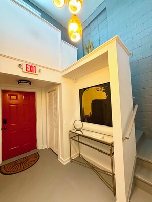 Un couloir avec un panneau indiquant la sortie, une porte rouge, un mur blanc orné d’un tableau abstrait noir et jaune, et un escalier.