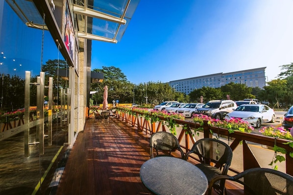 Ein Sitzbereich im Freien mit Tischen und Stühlen, einer Holzterrasse und im Hintergrund eine Ansicht auf geparkte Autos sowie ein Gebäude.