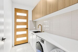A compact kitchen with a wooden door, white cabinetry, a built-in washing machine, and a sink with a gold faucet. - Indented Head Coastal Retreat | 5BR Escape (Indented Head)