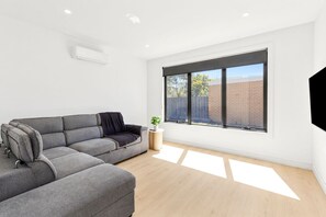 A modern living room with a grey sectional sofa, a small round side table, and a large window with a view of a brick wall and trees. - Indented Head Coastal Retreat | 5BR Escape (Indented Head)