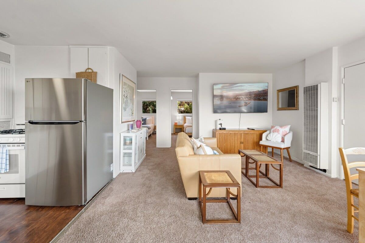 A modern kitchen with a stainless steel refrigerator, a dining area with a wooden table and chairs, and a living room with a beige sofa.