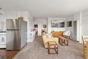 A modern kitchen with a stainless steel refrigerator, a dining area with a wooden table and chairs, and a living room with a beige sofa. - 2BD Ocean View | Private Deck + BBQ | Wellness (Summerland)