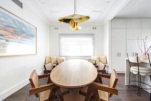 Uma sala de jantar com mesa redonda de madeira rodeada por cadeiras, luminária pendente e uma grande janela com persianas.