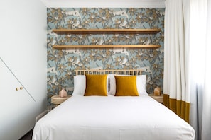 Cama com roupa de cama branca, duas almofadas mostarda e cabeceira com prateleira de madeira.