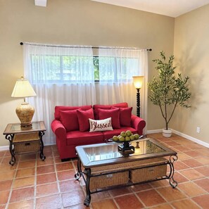A living room with a red sofa, a glass coffee table, a lamp, a potted plant, and a window with white curtains. - Family Tiki Bar, Hot Tub & Grill • 5 min SRQ (Sarasota)