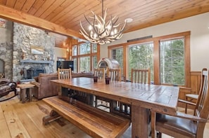 A wooden dining table with benches, a stone fireplace, and a large window with a view of trees. - Ski Trail to Door | Hot Tub, Games & Gym (Lincoln)