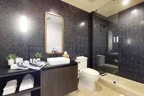 A modern bathroom with a dark speckled tile wall, a white sink, a toilet, and a glass-enclosed shower area. - 🌅 Rainey Street Retreat | Lake Views, Rooftop Pool & 31st Floor Skyline 🎸 (Austin)