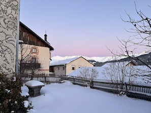 A snow-covered courtyard with a wooden fence, a building with a traditional design, and a mountain range in the background. - Turmhaus Giovanoli, (Tschlin) by Interhome (Tschlin)
