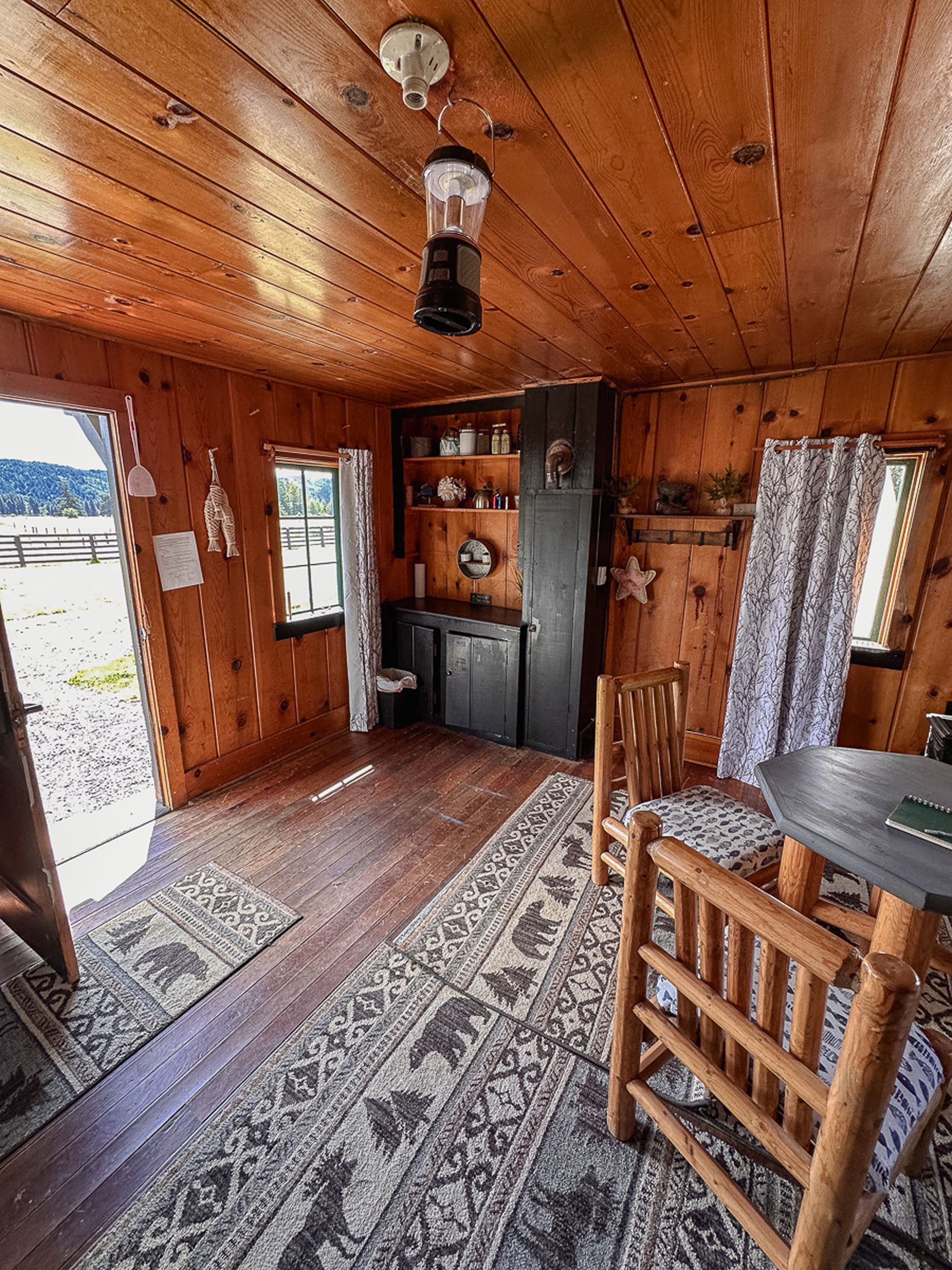 Ein Interieur einer Holzhütte mit Holzboden, einem Esstisch, einem Stuhl, einer Küchenzeile inklusive Kühlschrank und einem Fenster mit Vorhängen.