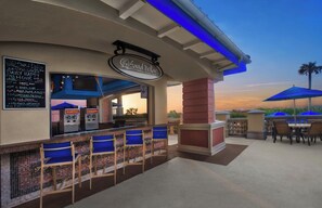 Eine Außenbar mit einem Schild, auf dem 'Sand Dollar Pool Bar' steht, Sitzgelegenheiten mit blauen Kissen und Blick auf den Sonnenuntergang.