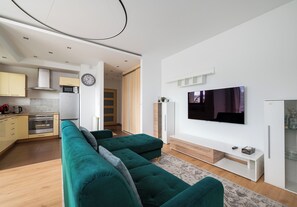 A modern living room with a large flat-screen TV, a teal sofa, and a kitchen in the background. - Olbrachta Cozy Nest Apartment (Warszawa)