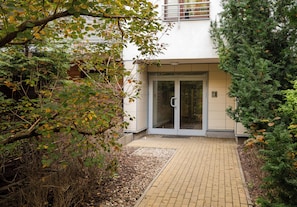 Una entrada de edificio con puerta de cristal, sendero pavimentado y follaje circundante.