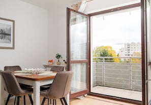 A dining area with a table and chairs, a window with a view of buildings and trees, and a balcony with a railing. - Olbrachta Cozy Nest Apartment (Warszawa)