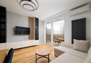 A modern living room with a flat-screen TV, a round wooden table, a sofa, and a balcony with a view of buildings. - Ursus Serene Apartment (Warszawa)