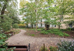 Ein Kiesweg führt durch einen Park mit Bänken, Bäumen und einem Gebäude im Hintergrund.