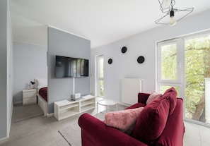 A modern living room with a flat-screen TV, a red sofa, a white coffee table, and a large window with a view of trees. - Olszowa 14 Riverside Apartment (Warszawa)