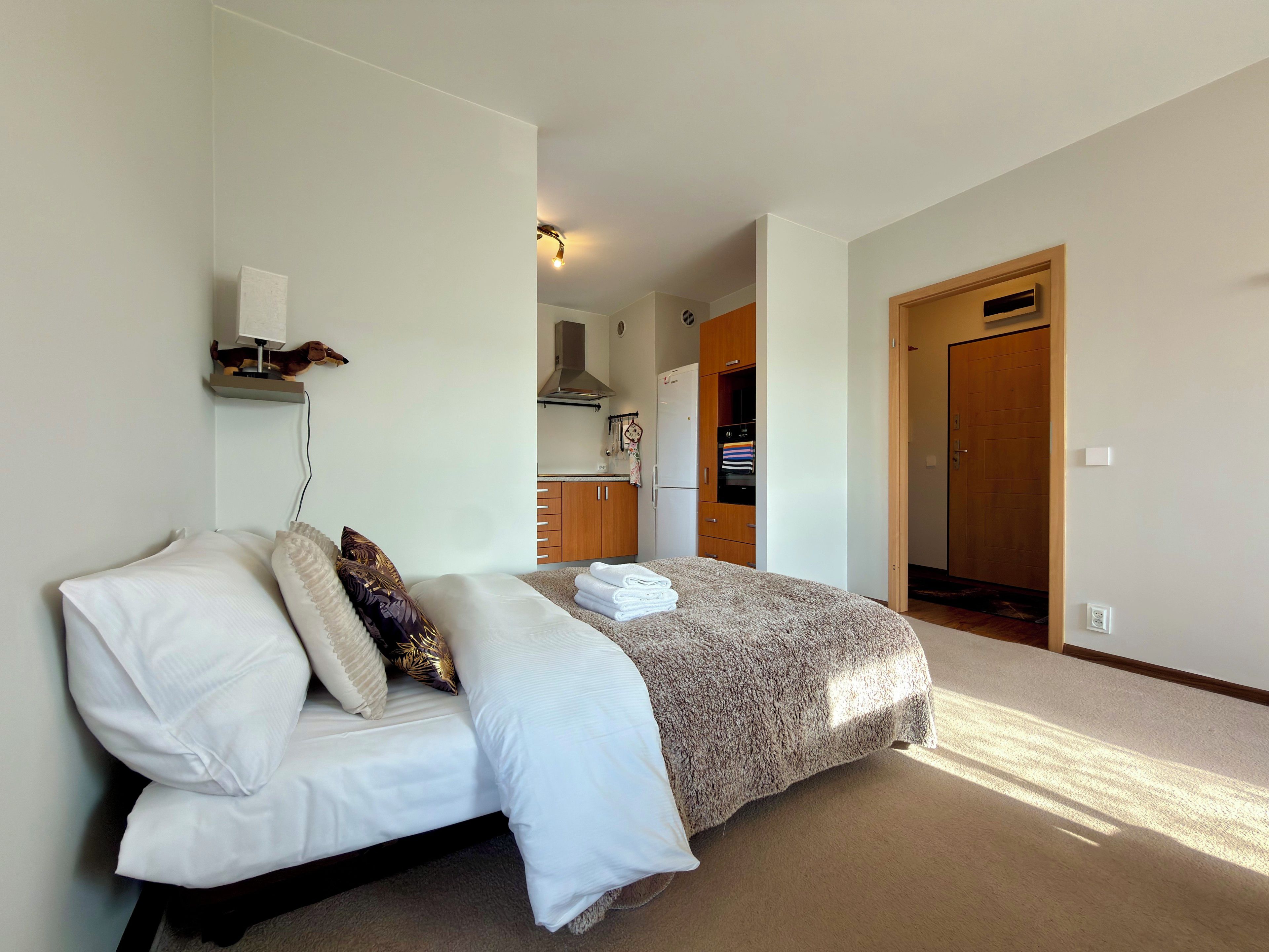 Une chambre à coucher comprenant un lit, une table de chevet, une étagère fixée au mur et une porte donnant sur un espace cuisine.
