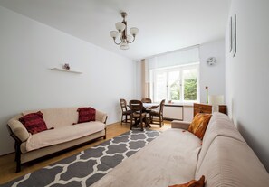 A well-lit room with a sofa, a dining table with chairs, a small window with a view, and a wall clock. - Saska Kępa Standard Apartment (Warszawa)