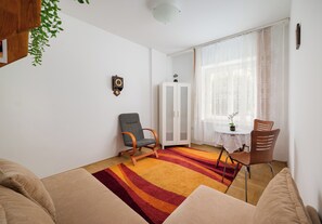 A living room with a sofa, a chair, a small table with a plant, a clock on the wall, and a window with sheer curtains. - Saska Kępa Standard Apartment (Warszawa)