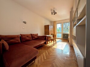 A modern living room with a brown sofa, wooden herringbone flooring, a dining table with chairs, and a view through large windows. - Przestrzenna Comfort Apartment (Wrocław)